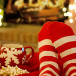 A pair of feet covered in red and white horizontal striped socks rest on a foot stool before a holiday fire next to a beverage and some treats